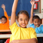 Three cheerful young primary school children indicating they know the answer with hands raised in class